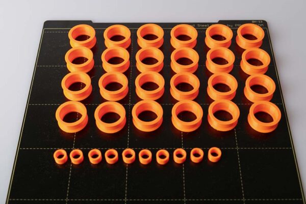Druckplatte des Prusa i3 MK3S auf der 24 Stück 3D gedruckte Rollen und 10 Stück 3D gedruckte Distanzstücke stehen. Alle Teile sind in orangen PETG Filament gedruckt.