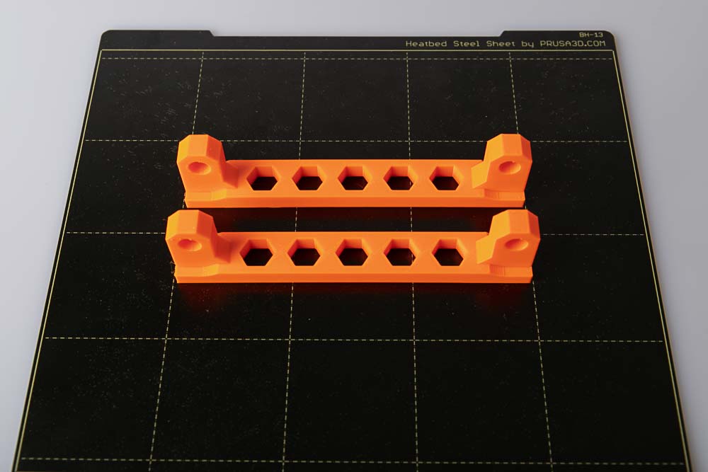 Print plate of the Prusa i3 MK3S on which two orange 3D printed rod mounts are placed. Details: Layer height 0.2 mm and Prusament PETG Prusa Orange