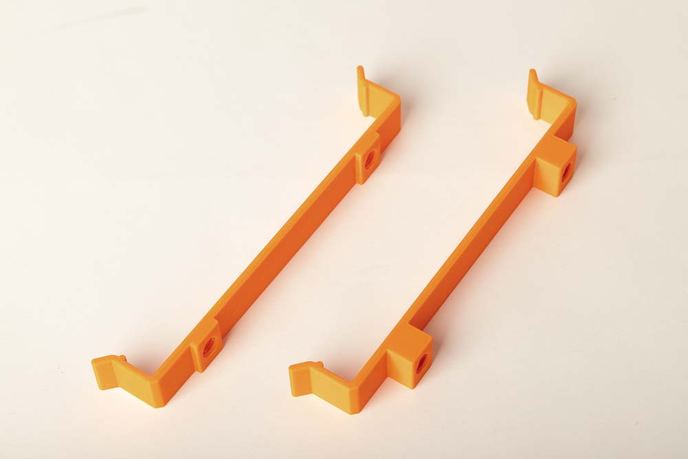 The two different 3D printed retaining clips for silica gel storage are located next to each other. The left bracket has a bar height of 3 mm and the right one has a bar height of 12 mm. The parts are printed in orange Prusament PETG with a layer thickness of 0.2 mm.