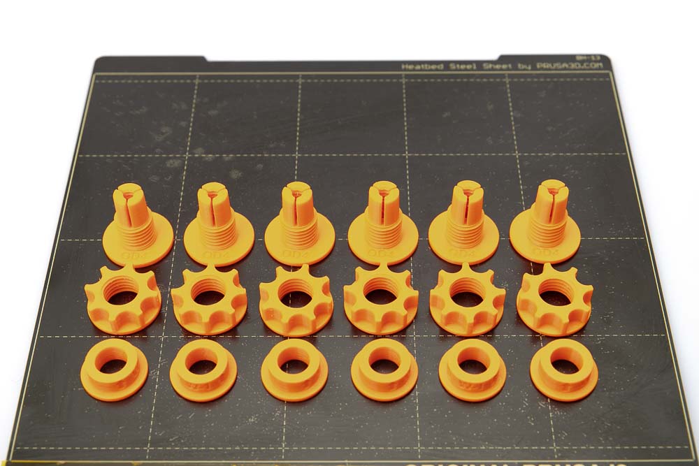 Printing plate of the Prusa i3 MK3S on which there are 6 pieces of 3D printed filament outlets, nuts and clamping rings. All parts are printed in orange PETG filament with a layer height of 0.2 mm.
