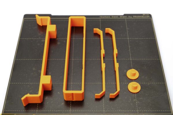Druckplatte des Prusa i3 MK3S auf dem eine 3D gedruckte Klammer H12, zwei Schrauben, eine Silica Gel Sack Halterung und zwei Seitenstützen liegen. Die 3D Druck Teile wurden in orangen PETG Filament mit einer Schichthöhe von 0,2 mm gedruckt.