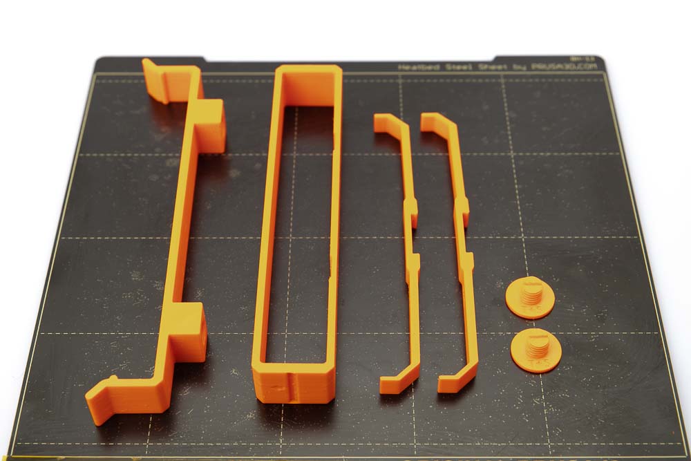 Print plate of the Prusa i3 MK3S on which there are placed a 3D printed clip_H12, two screws, a silica gel bag holder and two side supports. The 3D printed parts were printed in orange PETG filament with a layer height of 0.2 mm.