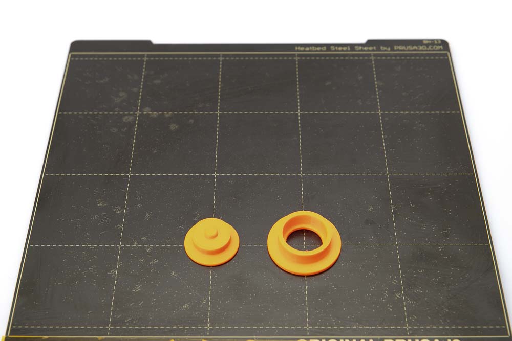 Druckplatte des Prusa i3 MK3S auf der die zwei Werkzeuge Einpresshilfe und Widerlager Auspresshilfe aufgelegt sind. Alle Teile sind in orangen Prusament PETG Filament mit einer Schichthöhe von 0,2 mm gedruckt