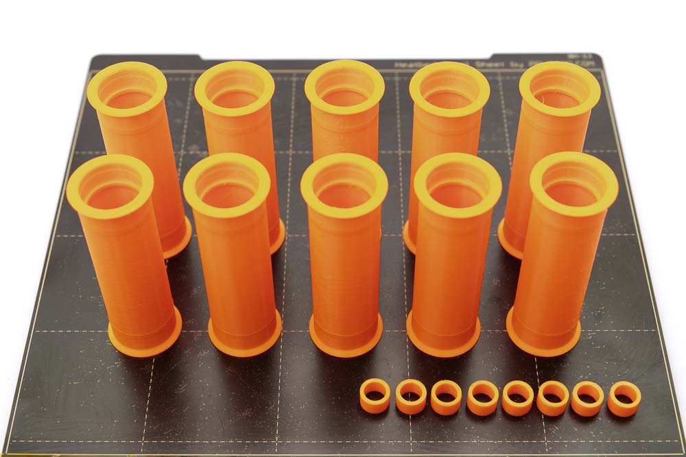 Druckplatte des Prusa i3 MK3S auf der 10 Stück 3D gedruckte breite Rollen mit Breite 80 mm und 8 Stück 3D gedruckte Distanzstücke stehen. Alle Teile sind in orangen PETG Filament gedruckt.