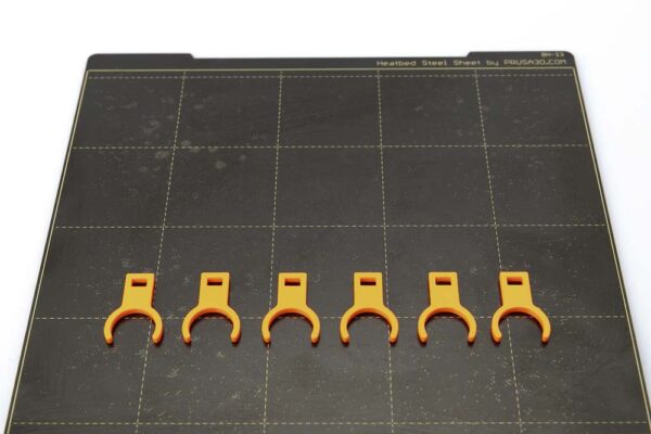 Druckplatte des Prusa i3 MK3S auf dem 6 Clips Variante A liegen. Die Clips wurden in orangen PETG Filament mit einer Schichthöhe von 0,2 mm gedruckt.