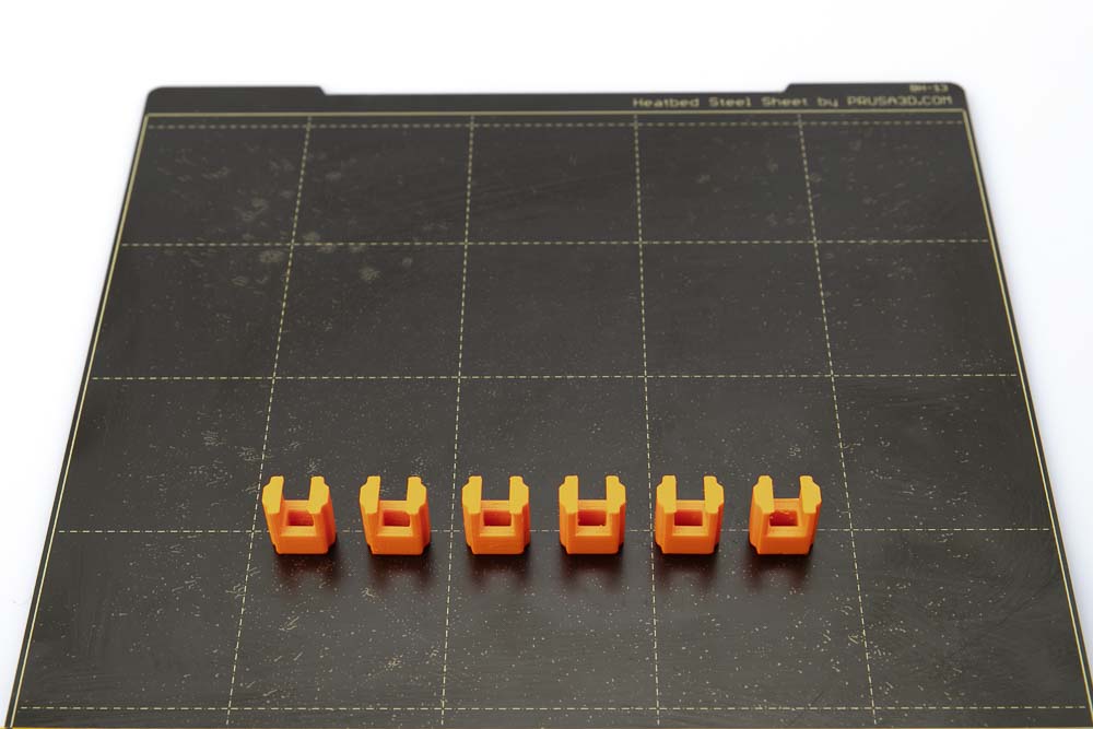 Druckplatte des Prusa i3 MK3S auf dem 6 Clips Variante B liegen. Die Clips wurden in orangen PETG Filament mit einer Schichthöhe von 0,2 mm gedruckt.