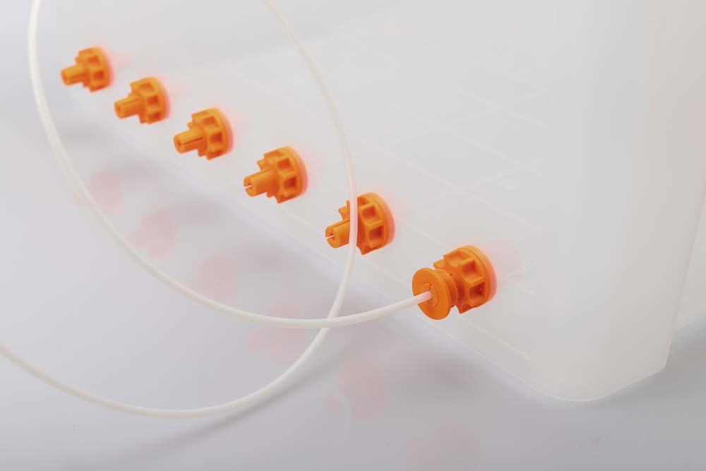 Luftdichte 50L Kunststoffbox mit sechs orangen Filament Durchführungen, alle mit einer 3D gedruckten Mutter gesichert. In der ersten Durchführung steckt ein langes (90 cm) Stück PTFE Schlauch welches mit einem 3D gedruckten Klemmring gesichert ist.