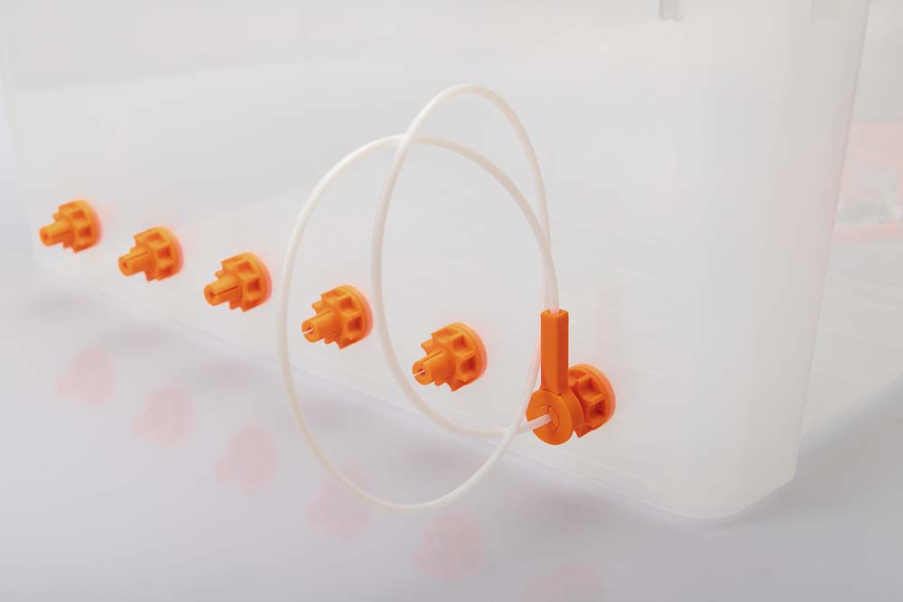 Luftdichte 50L Kunststoffbox mit sechs orangen Filament Durchführungen, alle mit einer 3D gedruckten Mutter gesichert. In der ersten Durchführung steckt ein langes (90 cm) Stück PTFE Schlauch welches mit einem 3D gedruckten Klemmring gesichert ist. Das Ende des Schlauches steckt in dem auf den Klemmring aufgeklipsten 3D gedruckten Stoppel der Variante B der Filament Box.
