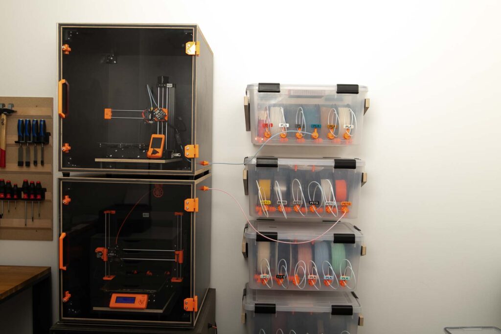 3D Druck Farm. Mehrere Filament Boxen auf Wandhalterungen und zwei 3D Drucker in 3D Drucker Gehäuse.