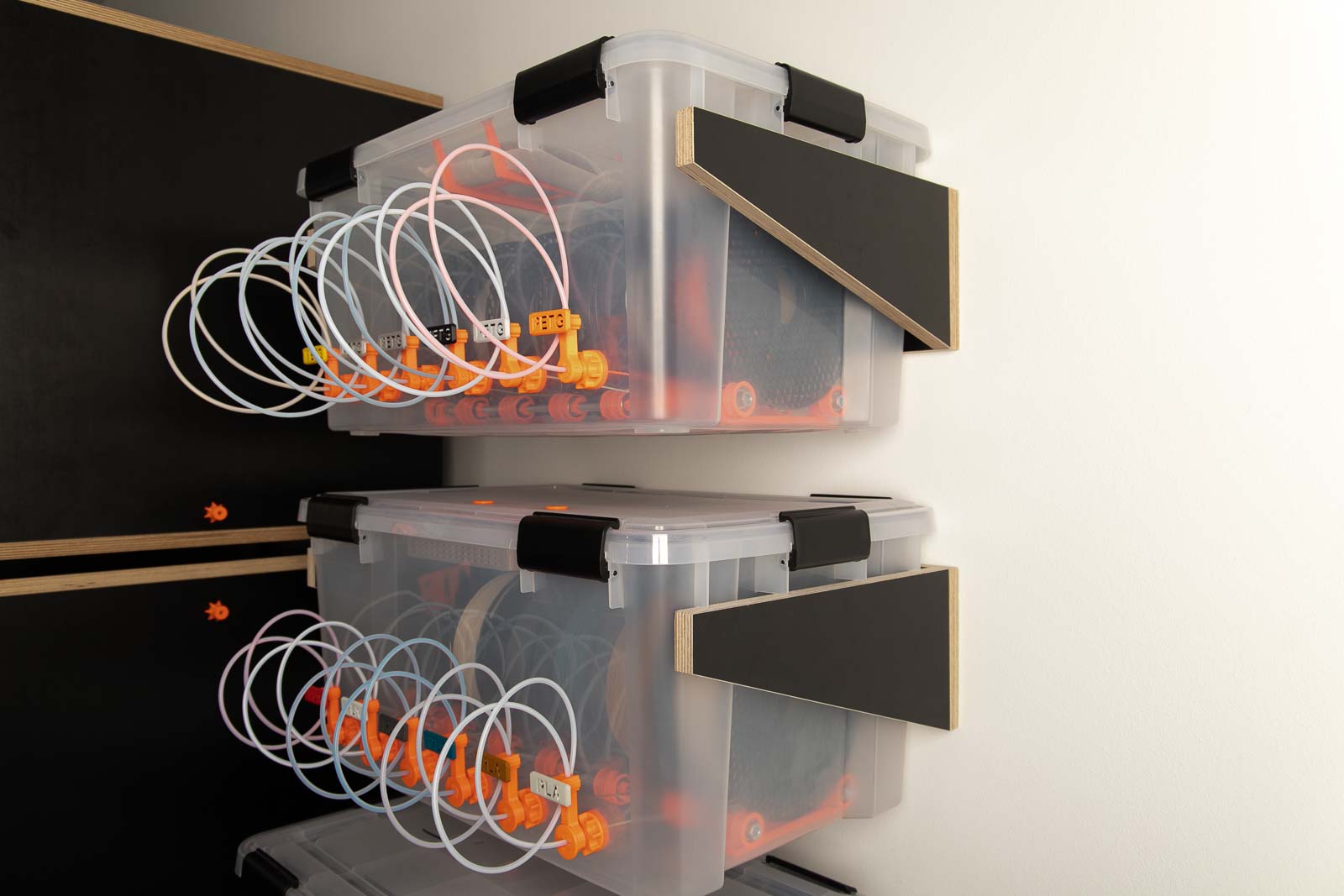 Zwei DIY Filament Boxen auf selbstgebauten Halterungen aus Holz. Im Hintergrund zwei gestapelte 3D Drucker Gehäuse mit Anschluss für das Filament.